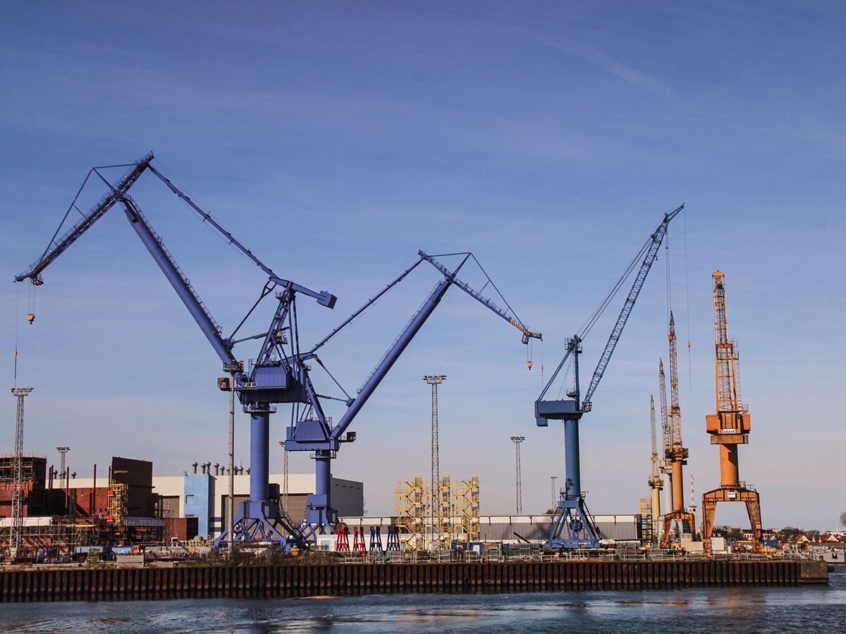 Blick auf einen Industriehafen mit mehreren großen blauen und orangefarbenen Kränen und Gebäuden unter blauem Himmel.