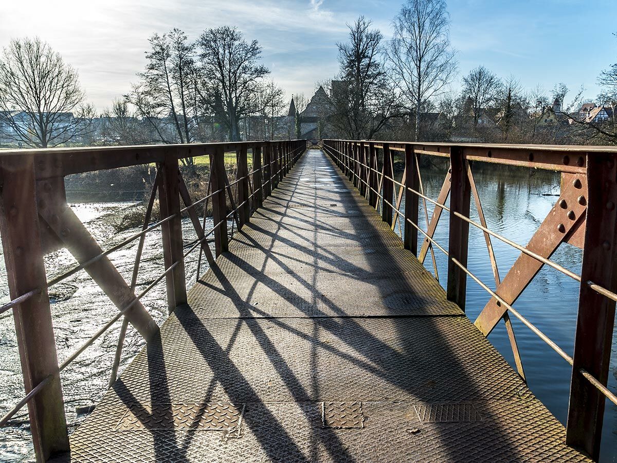 Eine lange, rostige Metallbrücke über einen Fluss mit kahlen Bäumen und einem Dorf im Hintergrund unter blauem Himmel mit Sonnenschein und Schatten.