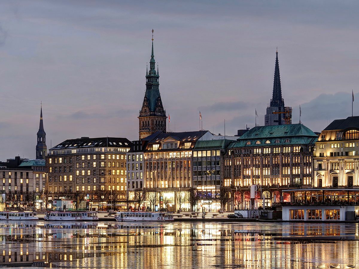 Abendlicher Blick auf die Hamburger Skyline mit beleuchteten Gebäuden, historischen Türmen und starken Lichtspiegelungen auf dem Wasser.