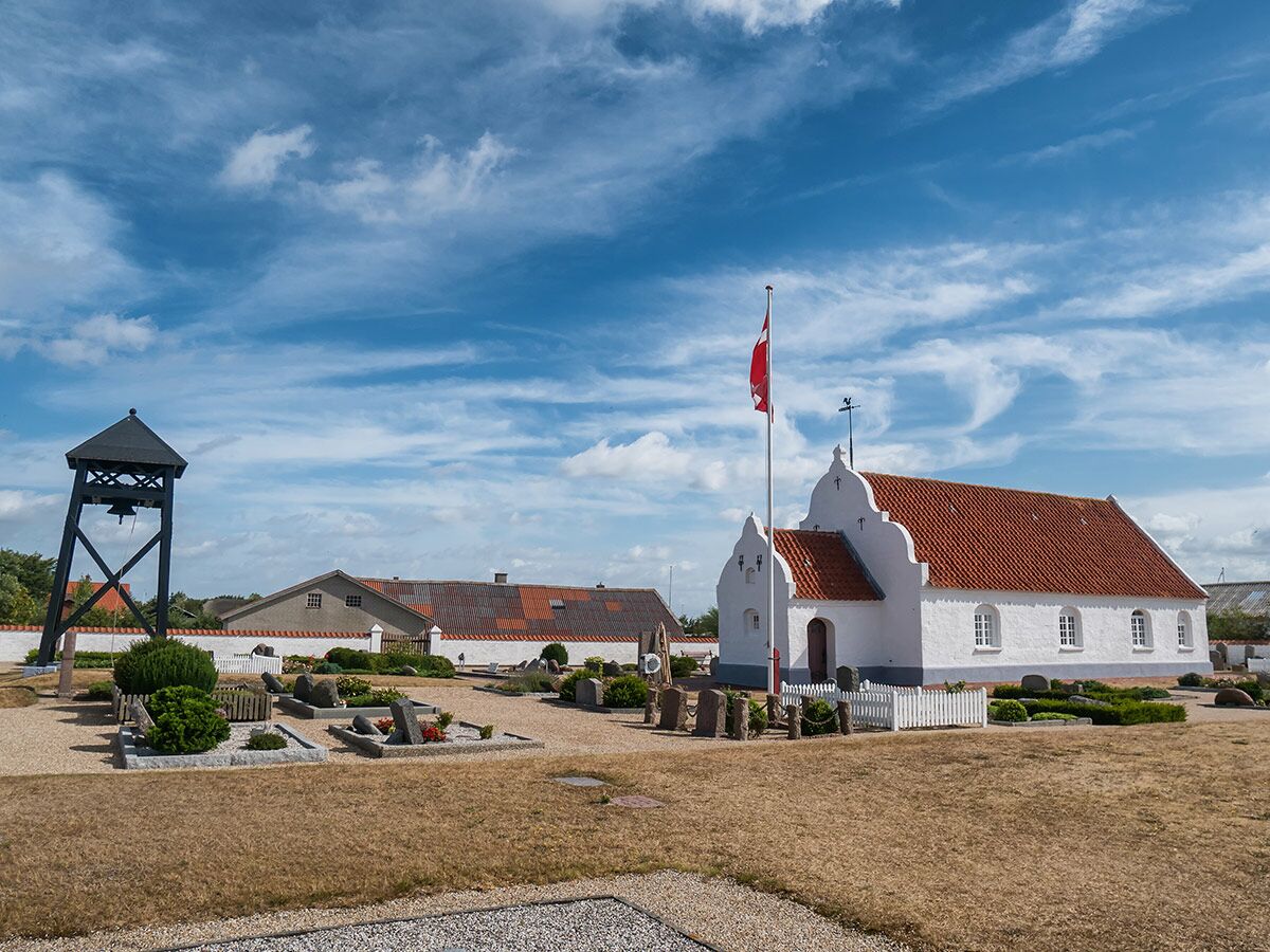 Weiße dänische Kirche mit rotem Dach, separatem Glockenturm und Friedhof unter blauem Himmel mit Wolken.