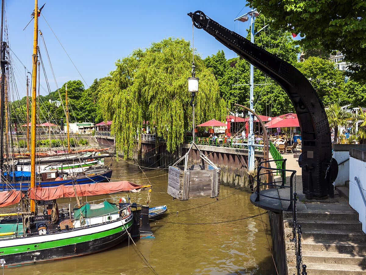 Idyllischer Hafen mit traditionellen Segelbooten, altem Kran, Café-Promenade und grünen Bäumen an sonnigem Tag.