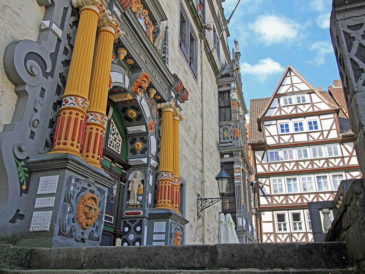 Blick auf historische Gebäude und ein Fachwerkhaus in einer deutschen Stadt, detailreiche Fassaden, Säulen und Verzierungen unter blauem Himmel.