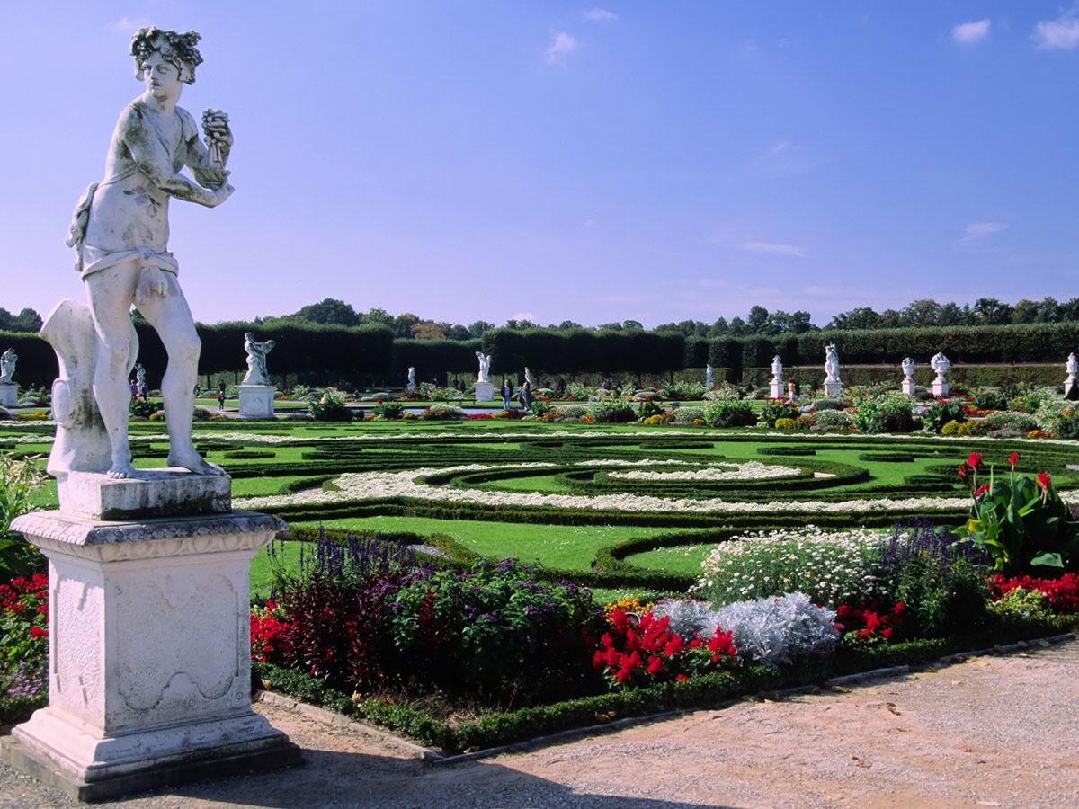 Prachtvoller Barockgarten mit einer weißen klassischen Statue im Vordergrund, umgeben von kunstvollen Blumenbeeten und Formschnitten.