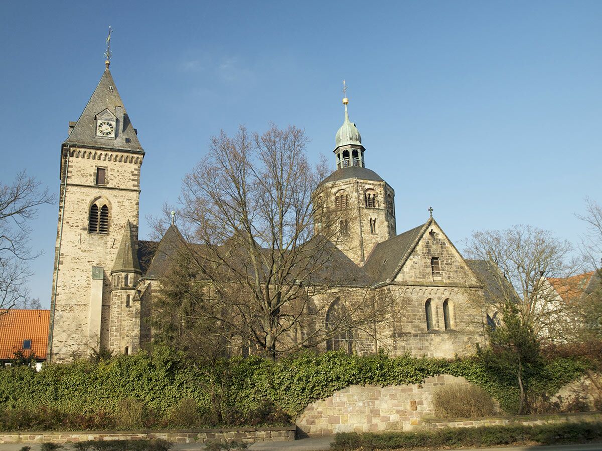 Historische, romanische Steinkirche mit zwei markanten Türmen unter blauem Himmel, umgeben von Bäumen und Efeu.
