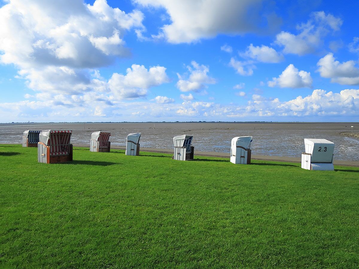 Reihe bunter Strandkörbe auf grüner Wiese vor dem Wattenmeer unter blauem Himmel mit Wolken an der Nordseeküste.