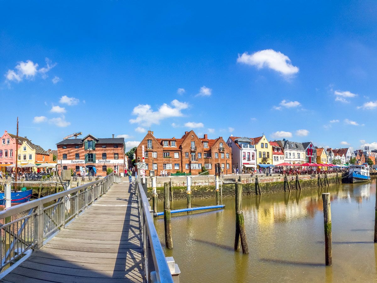Blick über einen Holzsteg auf bunte historische Häuser am Husumer Hafen unter blauem Himmel mit Wolken.