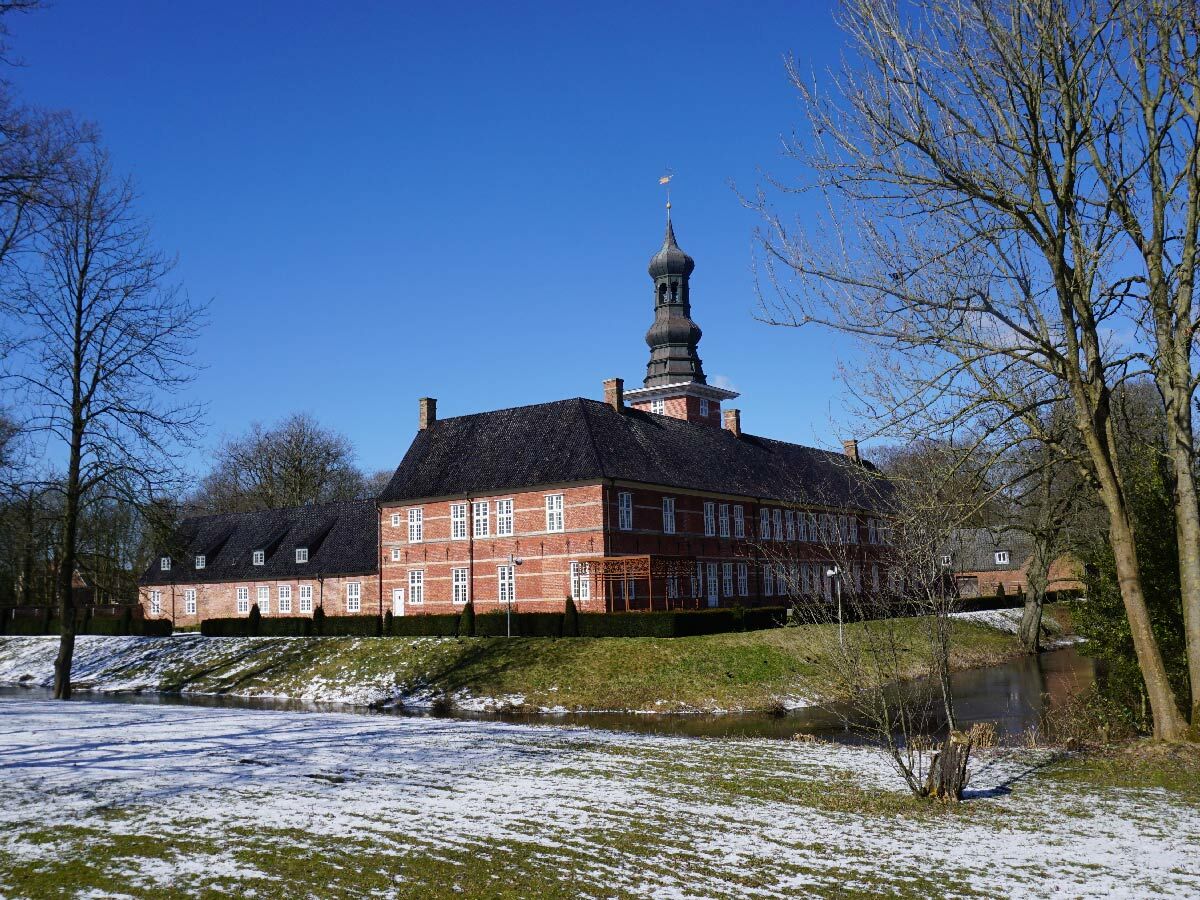 Historisches Ziegelgebäude mit markantem Turm und Schnee im Vordergrund, umgeben von Bäumen und einem Wassergraben unter blauem Himmel.