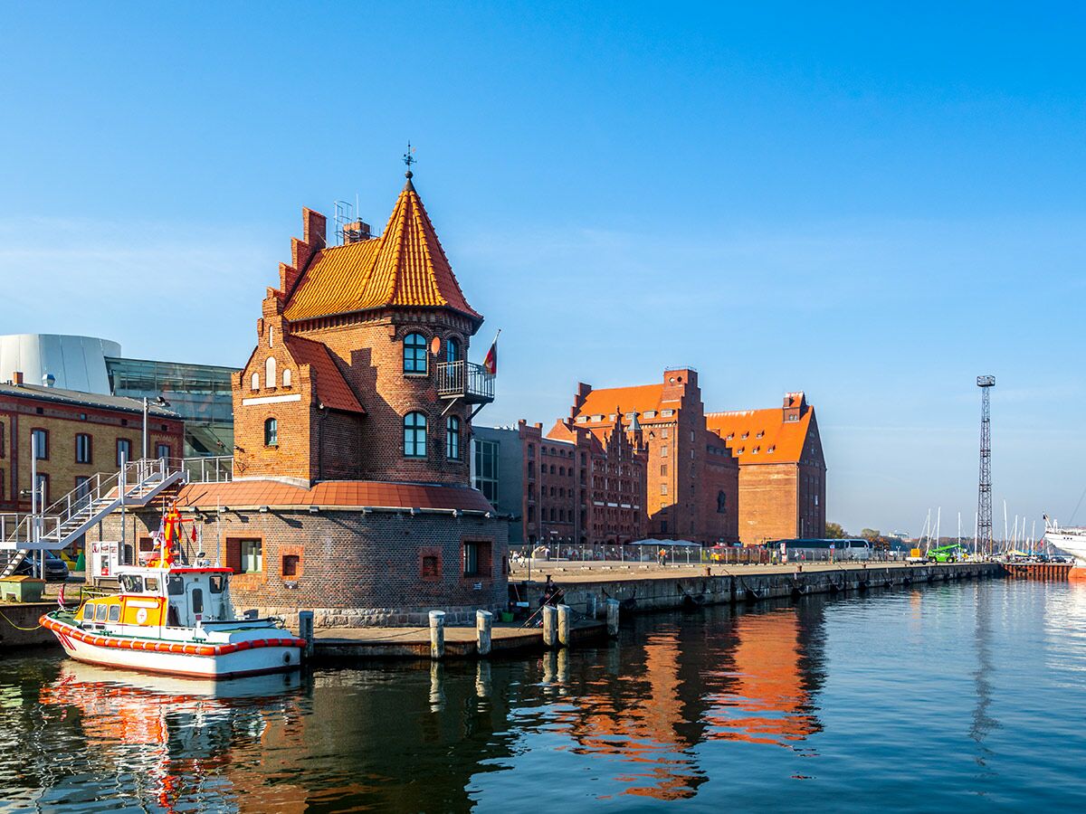 Historisches Backsteingebäude mit Turm und orangerotem Dach am sonnigen Hafen von Lübeck mit kleinem Boot im Vordergrund.