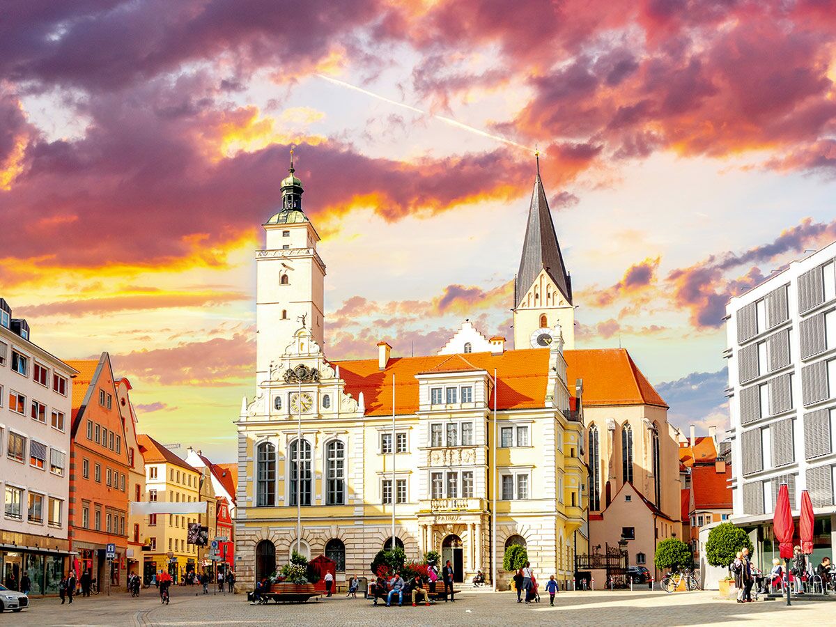 Belebter historischer Stadtplatz bei Sonnenuntergang mit Rathaus, Kirche, bunten Wolken und Menschen.