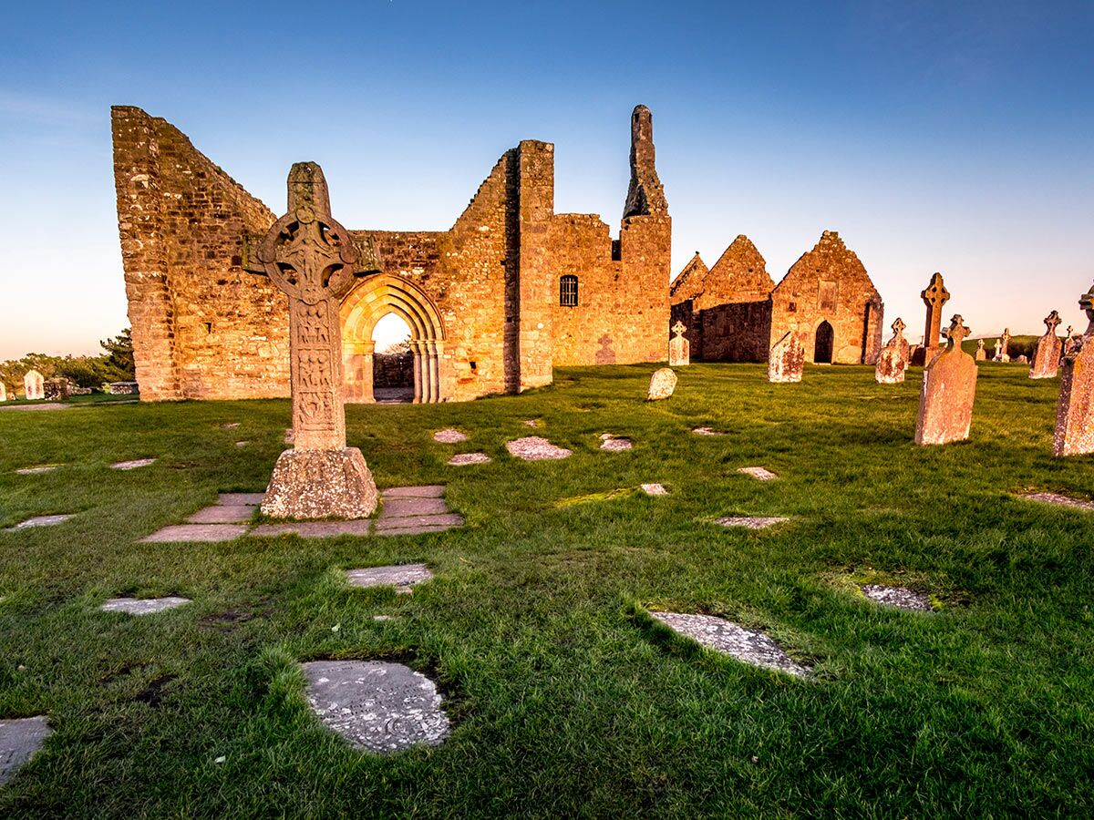 Historische Klosterruine mit keltischem Hochkreuz und alten Grabsteinen auf grünem Rasen bei Sonnenuntergang in Irland.