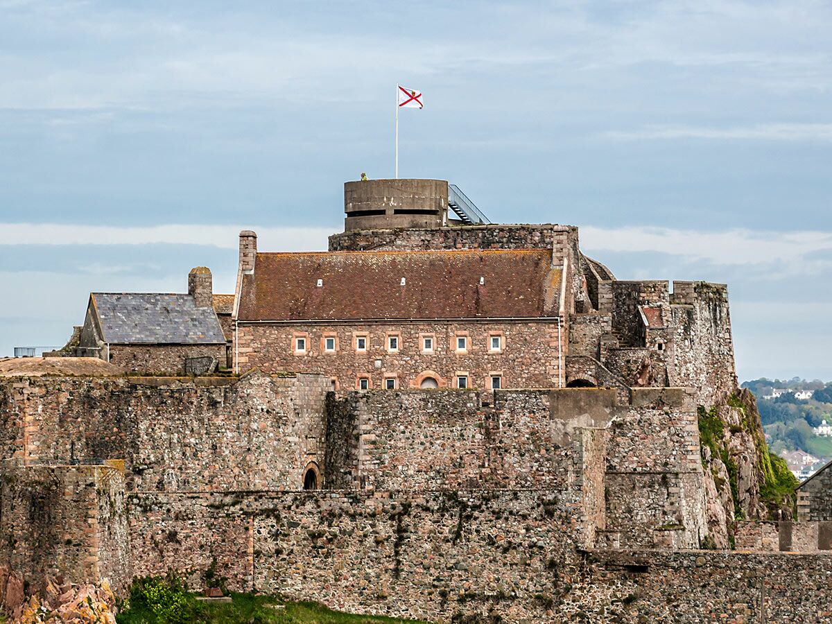 Ansicht der historischen Mont Orgueil Burg auf Jersey mit Steinmauern, Gebäuden, Bunker und gehisster Flagge unter blauem Himmel.