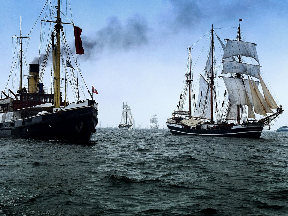 Ein historisches Dampfschiff und mehrere Großsegler fahren gemeinsam auf dem Meer unter einem bewölkten Himmel.