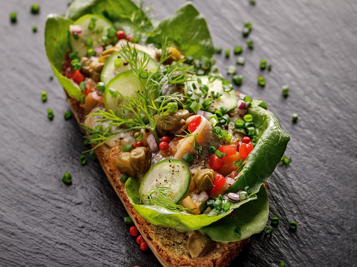 Frisch belegtes Brot mit Anchovis, Gurken, Dill, Kapern und roten Pfefferbeeren auf dunklem Schieferhintergrund.