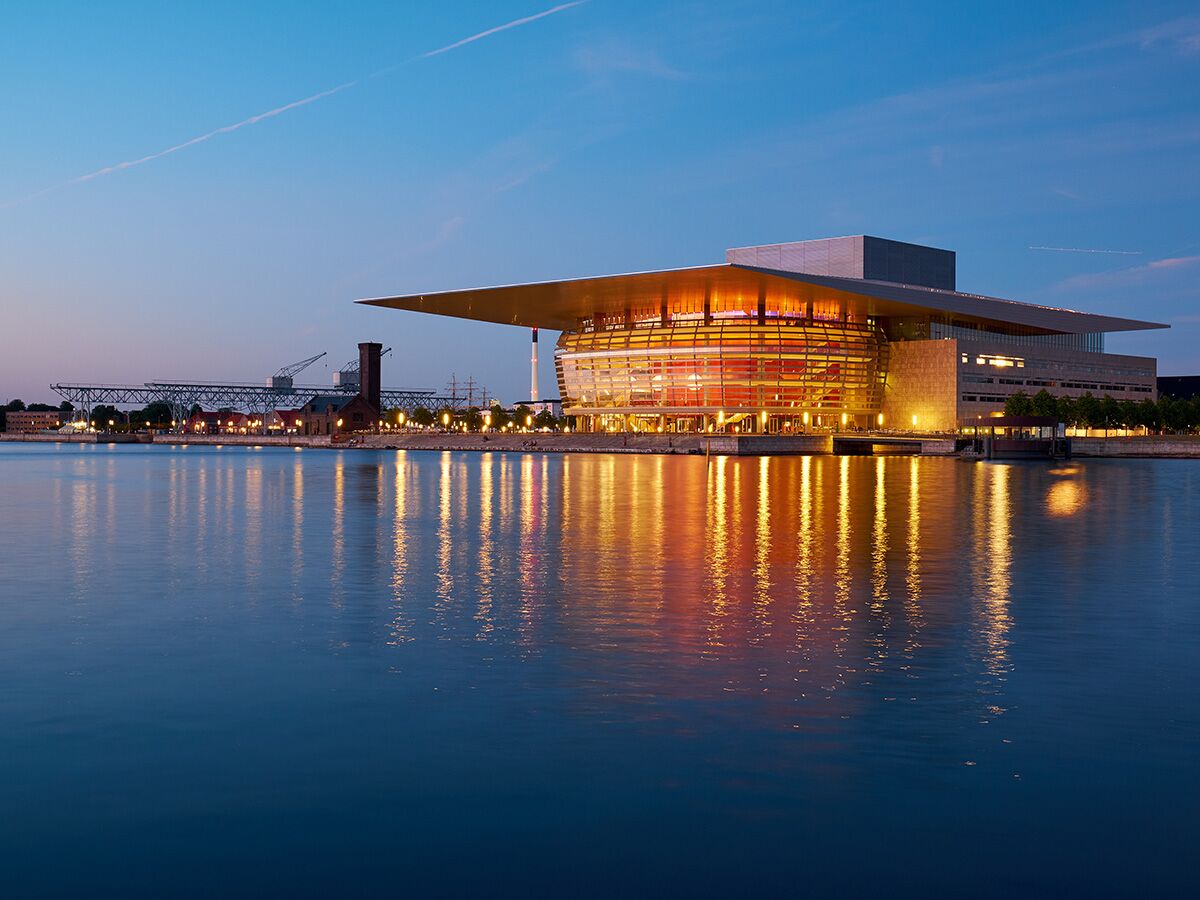 Kopenhagener Opernhaus bei Dämmerung, beleuchtet und im Wasser reflektiert, mit Hafenanlagen und Brücke im Hintergrund.