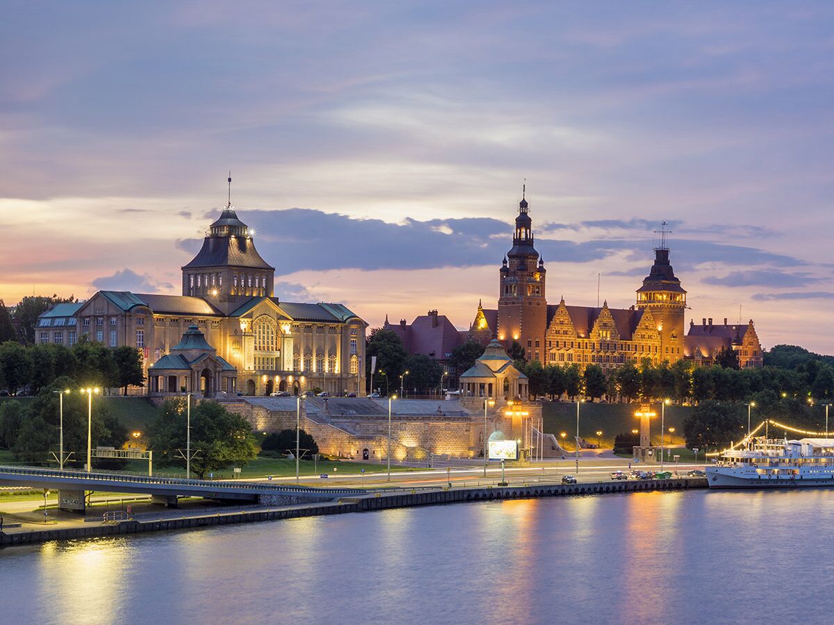 Panoramablick auf die beleuchtete Haken-Terrasse in Szczecin mit historischen Gebäuden, Fluss und Promenade bei Dämmerung.