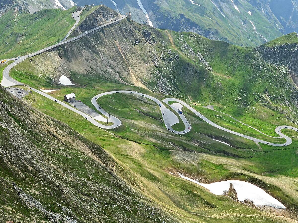 Spektakuläre Serpentinenstraße mit Brücke schlängelt sich durch grüne Alpenlandschaft mit Autos und Berghütte.