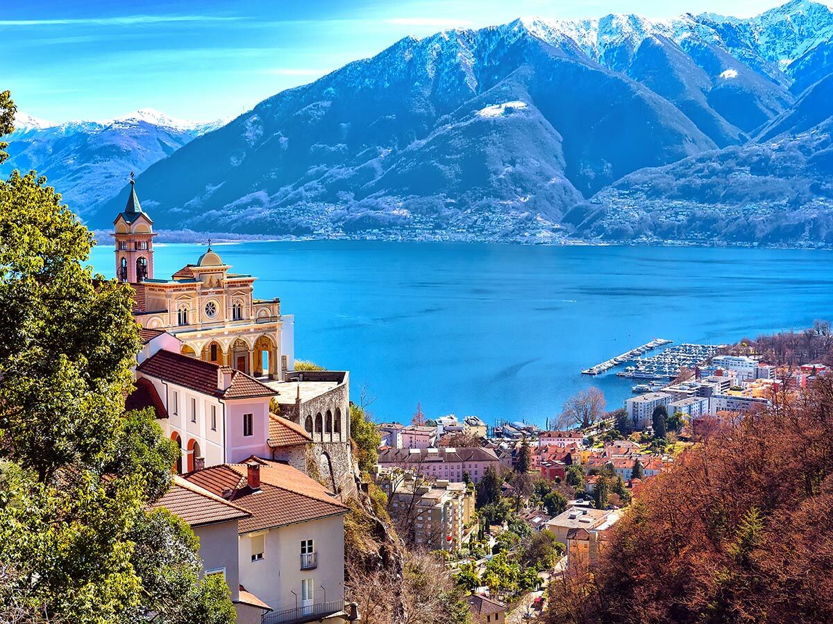 Panoramablick auf Locarno mit der Madonna del Sasso, dem Lago Maggiore und schneebedeckten Alpen unter blauem Himmel.