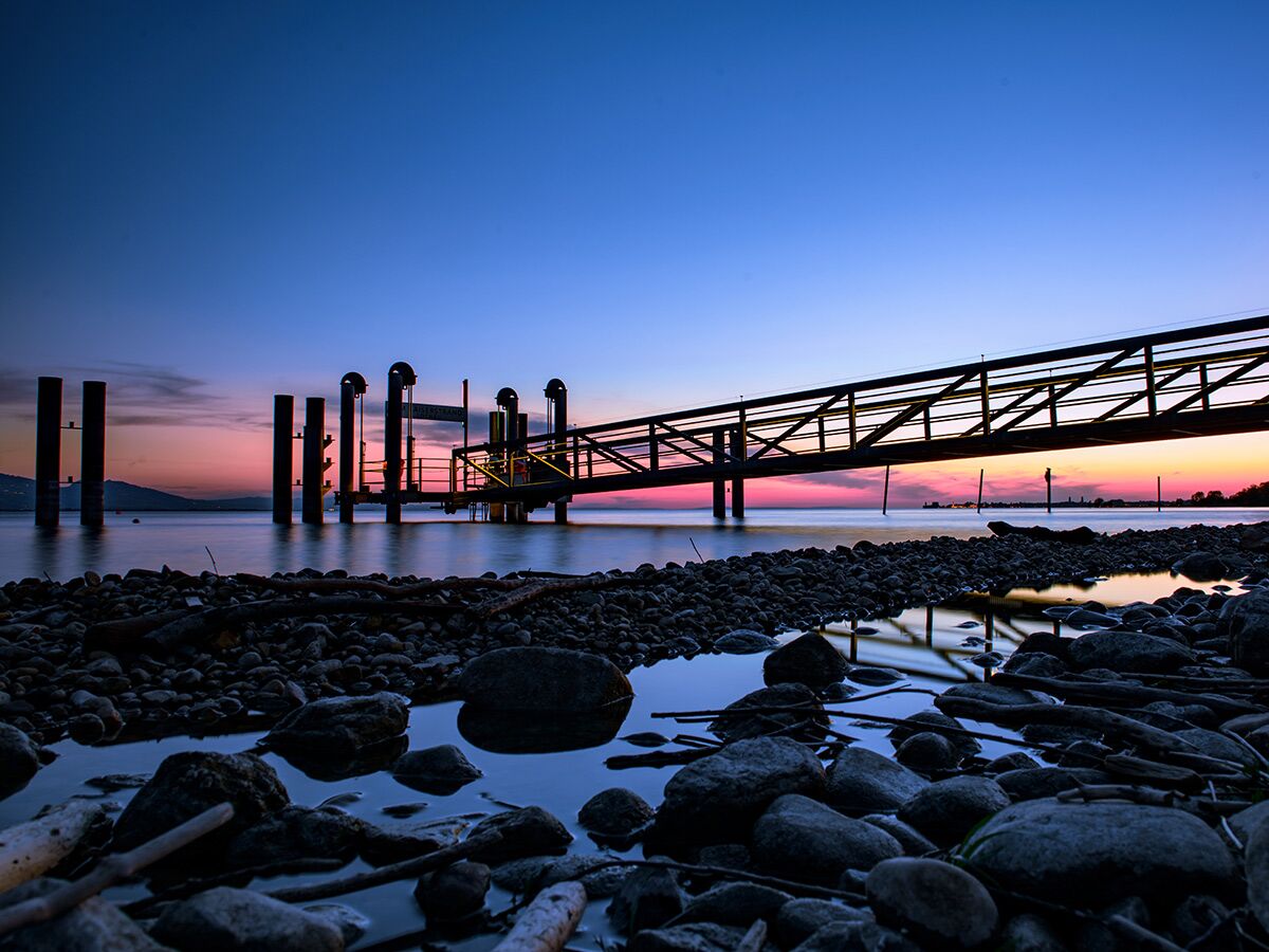 Uferlandschaft bei Sonnenuntergang mit einem langen Pier, der ins Wasser ragt, und Felsen im Vordergrund, Himmel blau-rosa.
