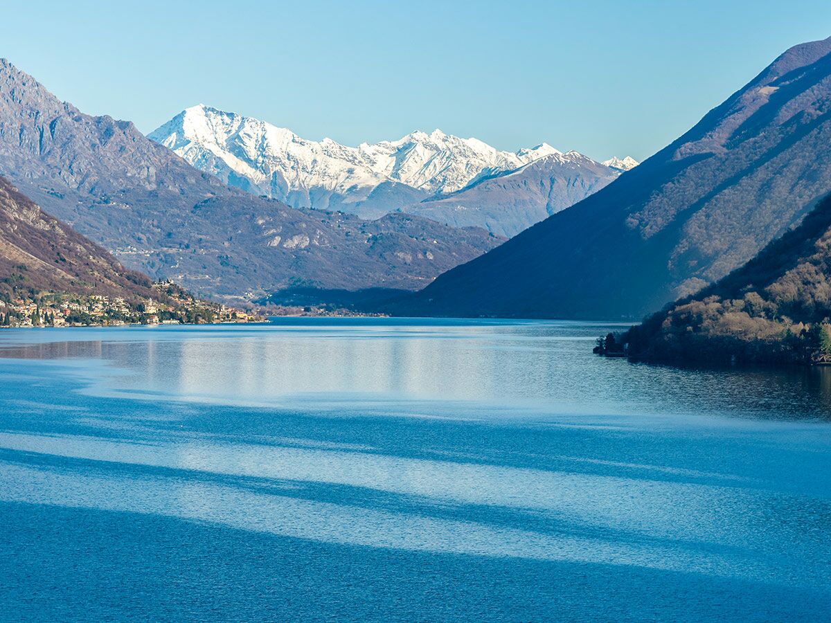 Malerischer Bergsee mit schneebedeckten Alpen und einem Dorf am Ufer unter blauem Himmel, ruhiges Wasser.