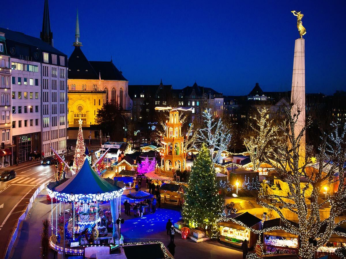 Beleuchteter Weihnachtsmarkt bei Nacht in Luxemburg Stadt mit Karussell, Weihnachtsbaum, Ständen und Gëlle Fra Denkmal.