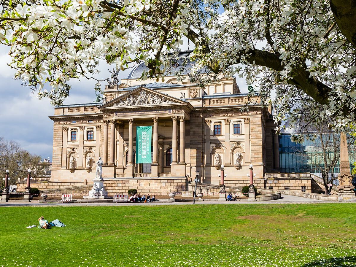 Das Hessische Staatstheater Wiesbaden im Frühling mit blühenden Magnolienbäumen und Menschen auf dem Rasen.