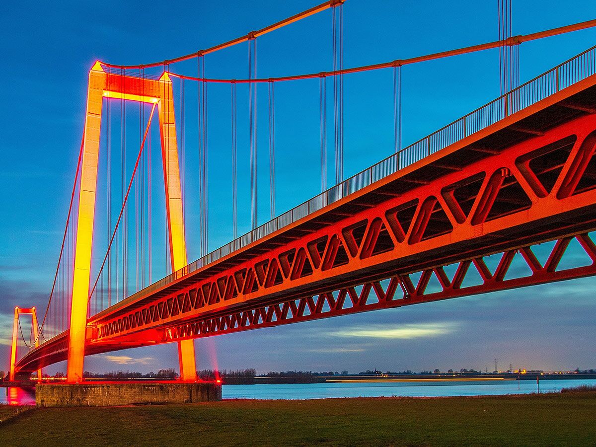 Orange beleuchtete Hängebrücke bei Dämmerung über blauem Fluss mit Reflexionen und dramatischem Himmel.
