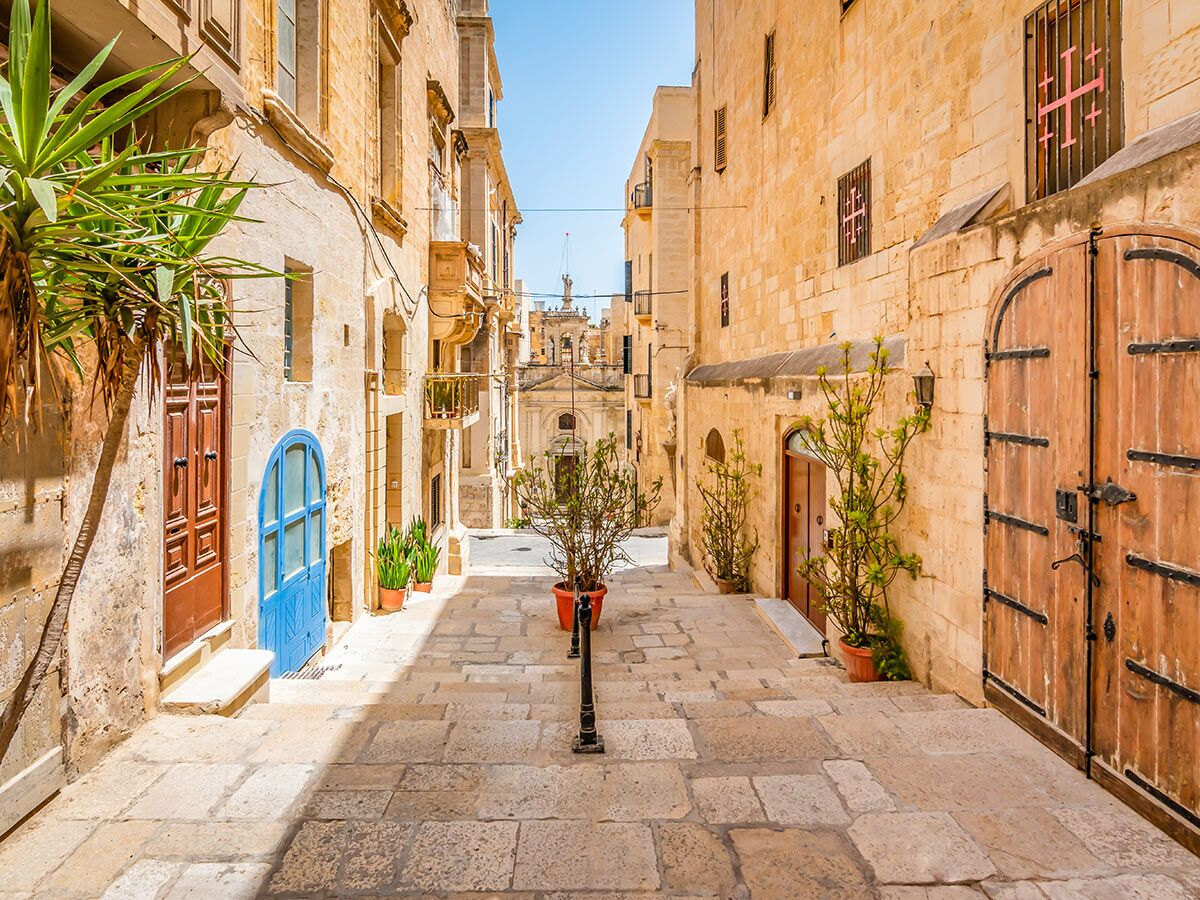 Schmale, sonnige Gasse in Vallettas Altstadt, Malta, mit historischen Steingebäuden, bunten Türen und Pflanzen.