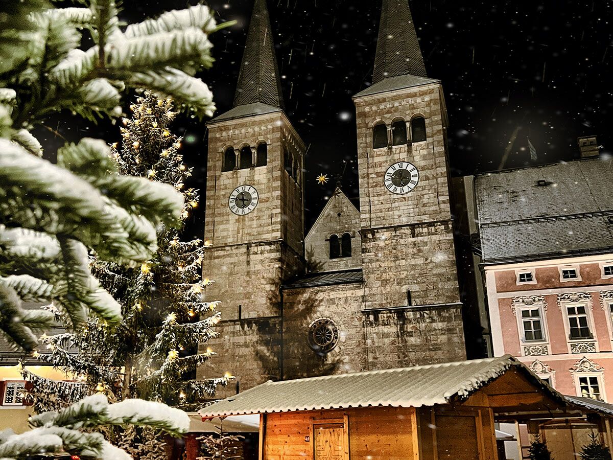 Verschneiter Weihnachtsmarkt bei Nacht mit beleuchtetem Christbaum, zwei Kirchtürmen und fallendem Schnee.