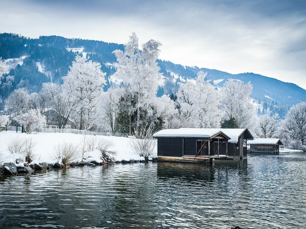Winterlandschaft am See mit hölzernen Bootshäusern, raureifbedeckten Bäumen und schneebedeckten Bergen im Hintergrund.