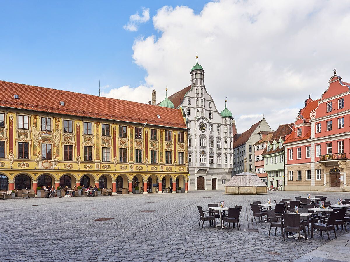 Lebhafter Marktplatz in Landsberg am Lech mit historischen Gebäuden, Außengastronomie und Brunnen unter blauem Himmel.