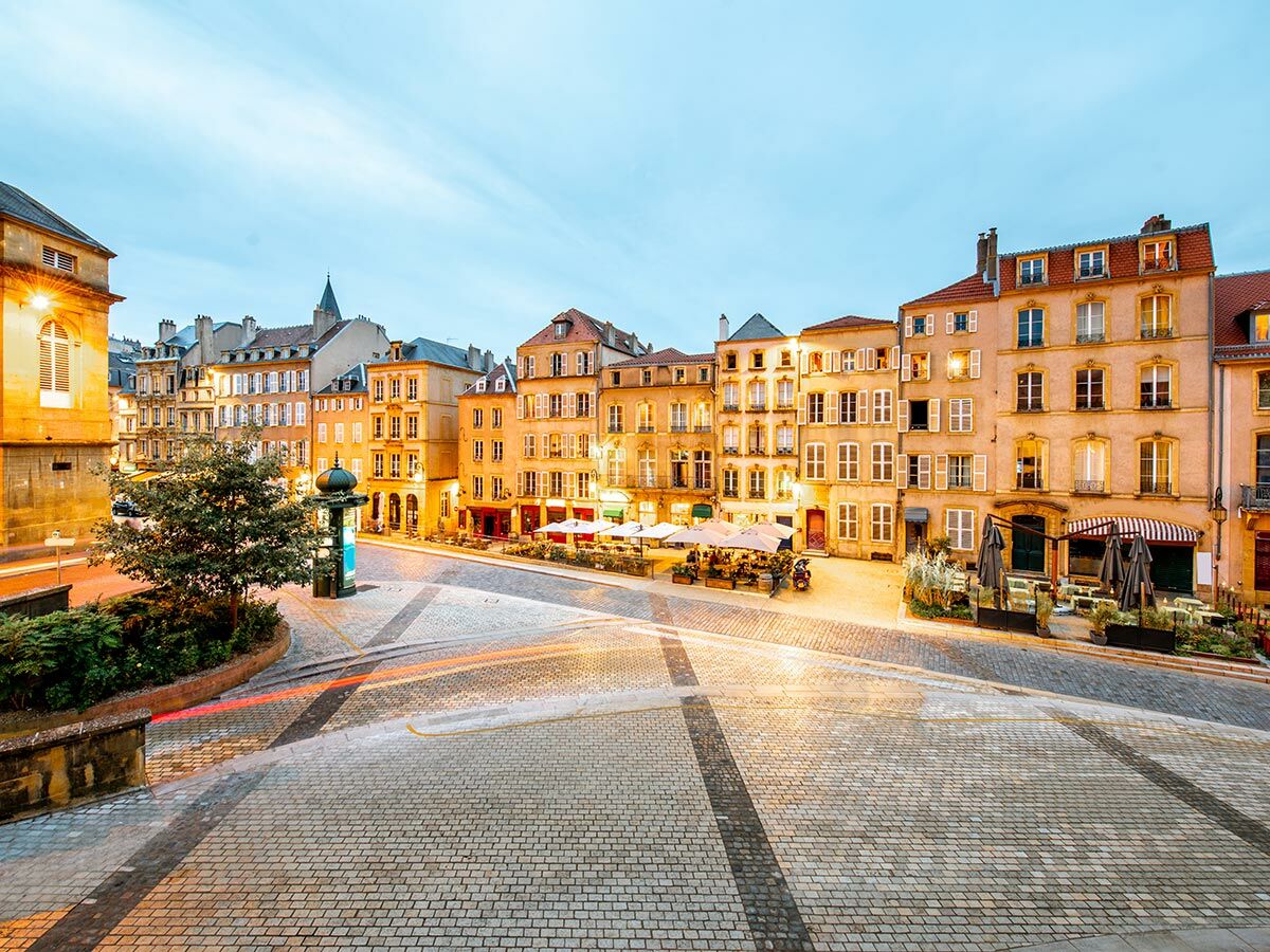 Abendliche Szene eines europäischen Stadtplatzes mit beleuchteten historischen Gebäuden, Kopfsteinpflaster, Restaurants und Lichtspuren von Fahrzeugen.