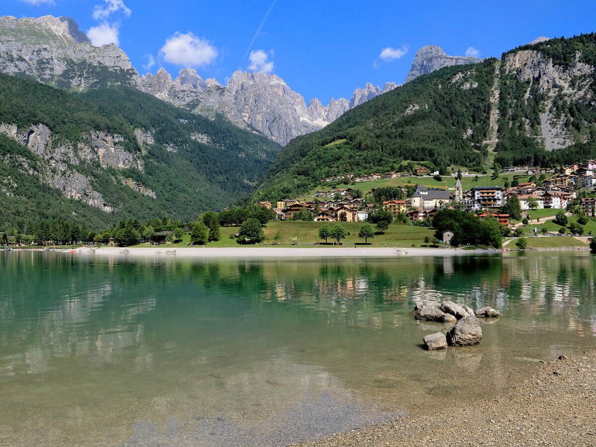 Malerischer Molvenosee in Trentino, umgeben von einem Bergdorf und den majestätischen Dolomiten, bei Sonnenschein.