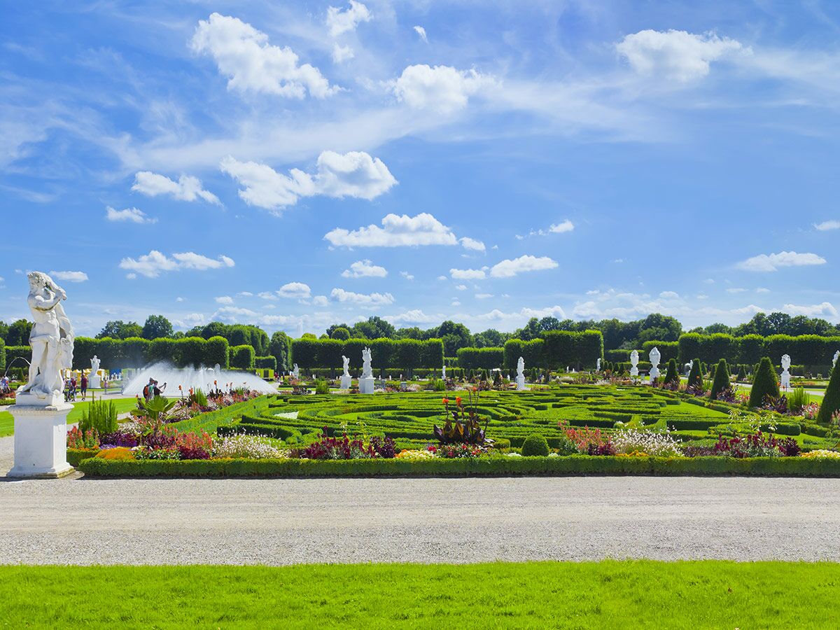 Großer, gepflegter Barockgarten mit kunstvoll geschnittenen Hecken, farbigen Blumenbeeten, Statuen und einem Springbrunnen.