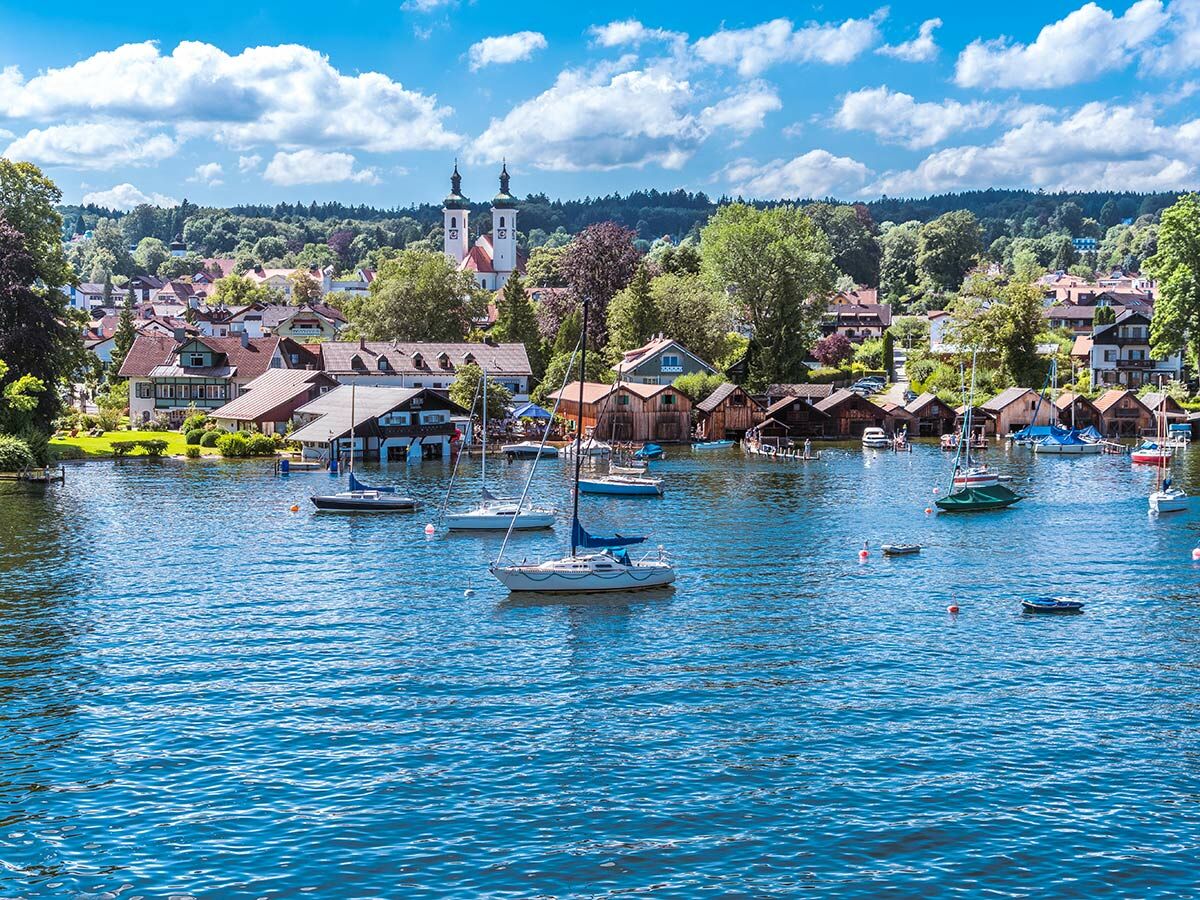 Malerisches bayerisches Dorf mit Boathäusern und Kirche am Seeufer. Segelboote schwimmen im blauen Wasser unter klarem Sommerhimmel.