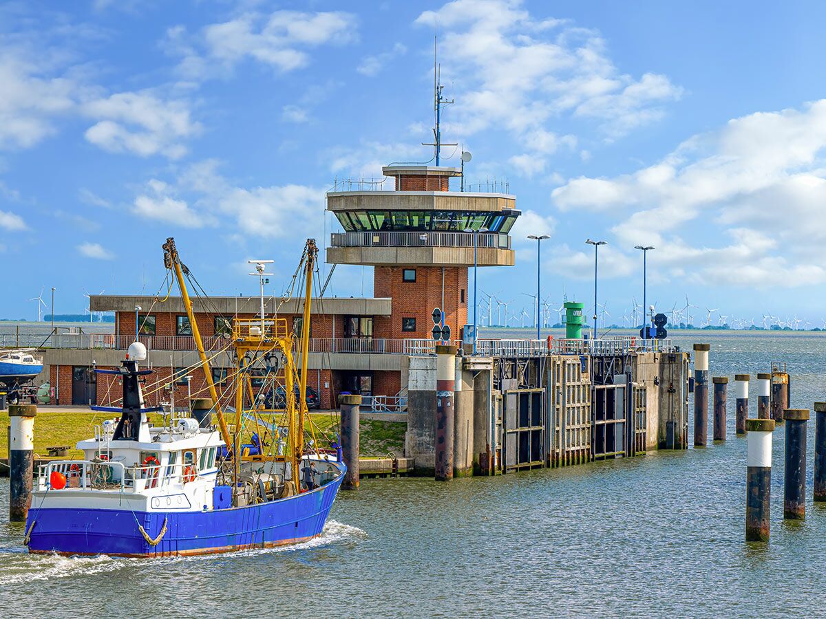 Blaues Fischerboot fährt aus dem Hafen mit Kontrollturm, Pier und Windkraftanlage im Hintergrund.