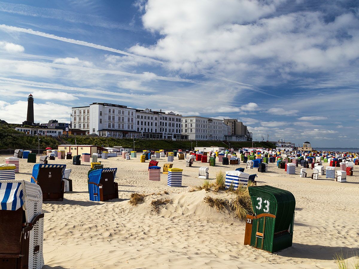 Ein sonniger Strand mit vielen bunten Strandkörben, einem Leuchtturm und einem großen weißen Hotel im Hintergrund.