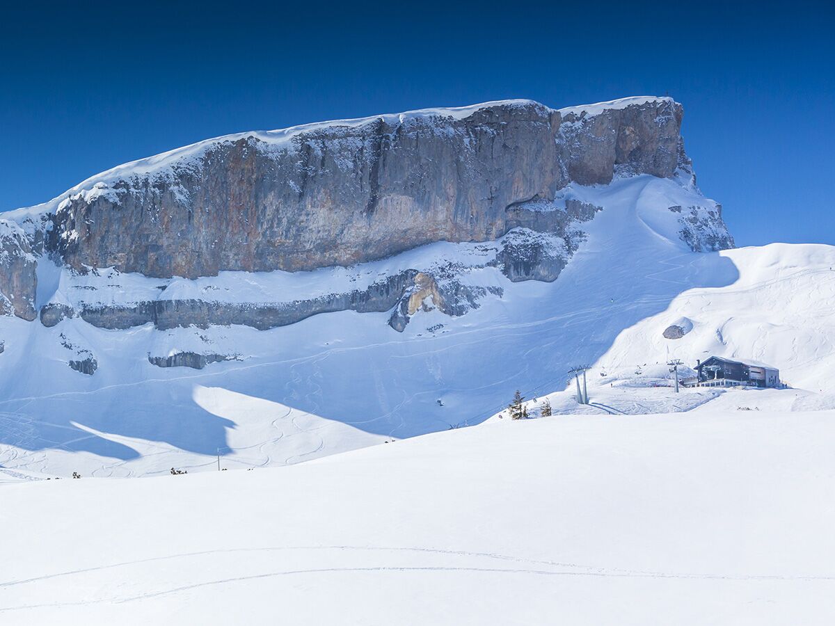 Winterliche Berglandschaft mit hoher Felswand, schneebedeckten Skipisten, Skiliften und einer Berghütte unter blauem Himmel.
