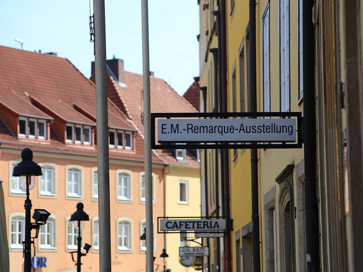 Ein Schild "E.M.-Remarque-Ausstellung" hängt an einem gelben Gebäude, im Hintergrund alte Häuser mit roten Dächern und weiteren Schildern.