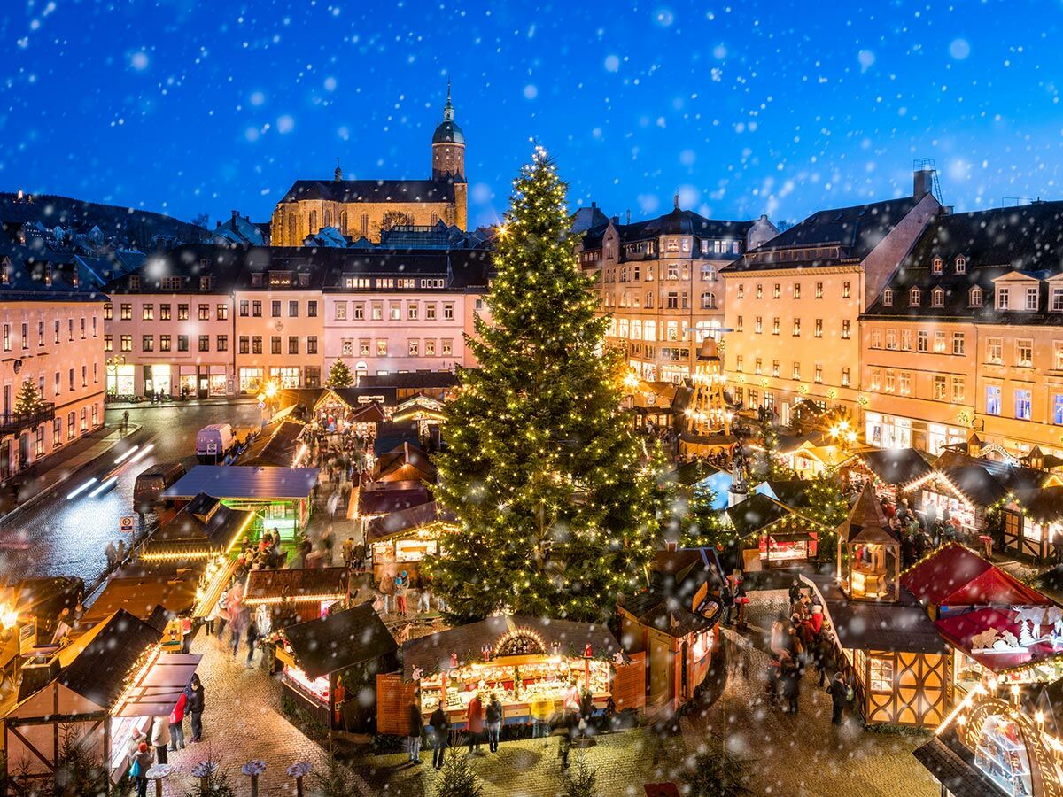 Vogelperspektive auf einen beleuchteten Weihnachtsmarkt in einer historischen Stadt bei Schneefall am Abend, mit großem Tannenbaum und geschäftigem Treiben.