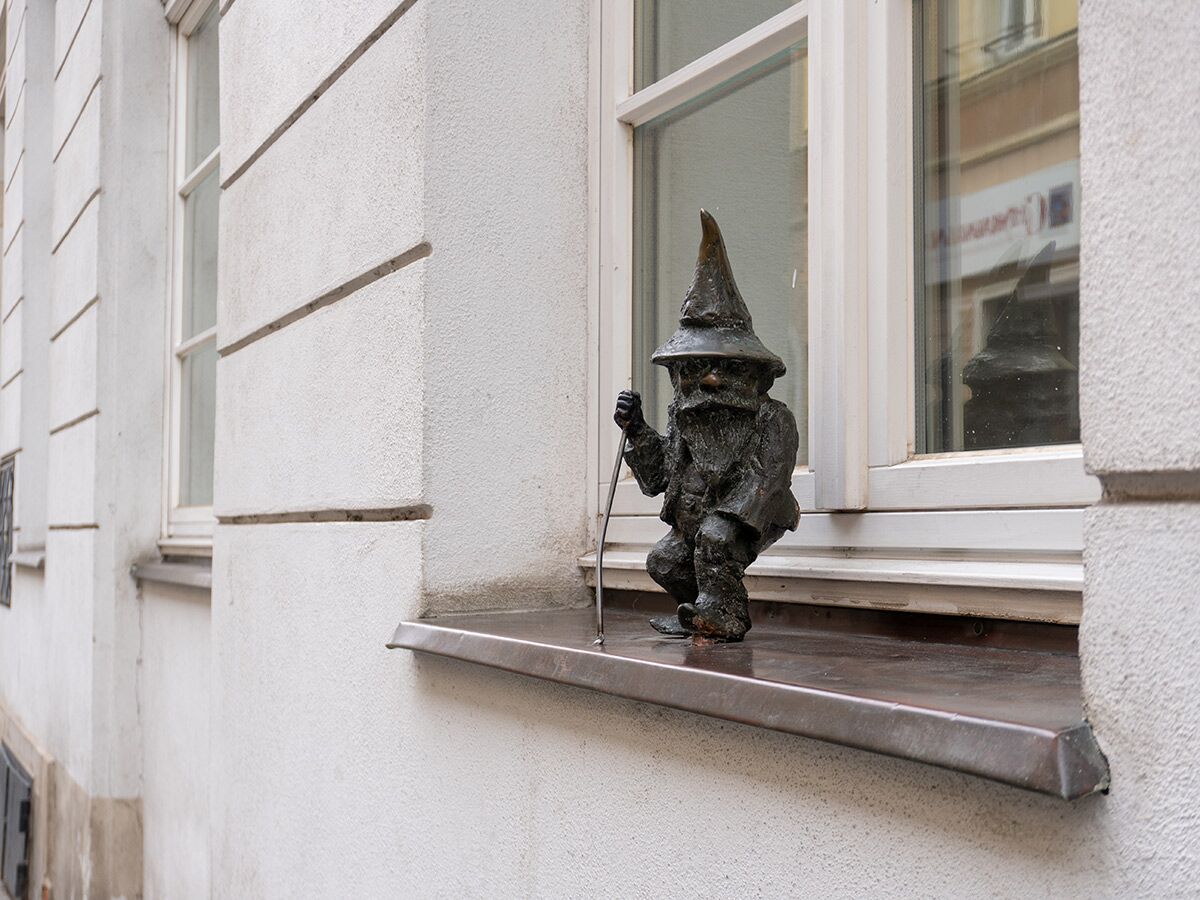 Kleine Bronzestatue eines Zwergs mit Spitzhut und Bart, der auf einer Fensterbank eines weißen Gebäudes sitzt.