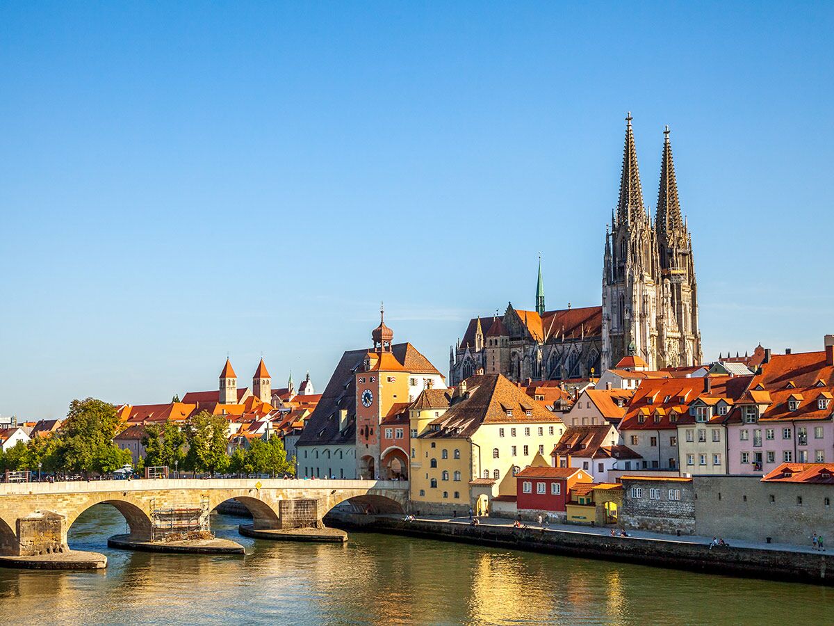 Panoramablick auf Regensburg mit dem Dom, Steinerner Brücke und bunten Häusern am Ufer der Donau unter blauem Himmel.