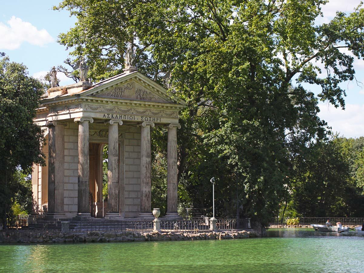Klassischer Tempel des Äskulap in der Villa Borghese, Rom, umgeben von Bäumen und einem grünen See mit Booten.