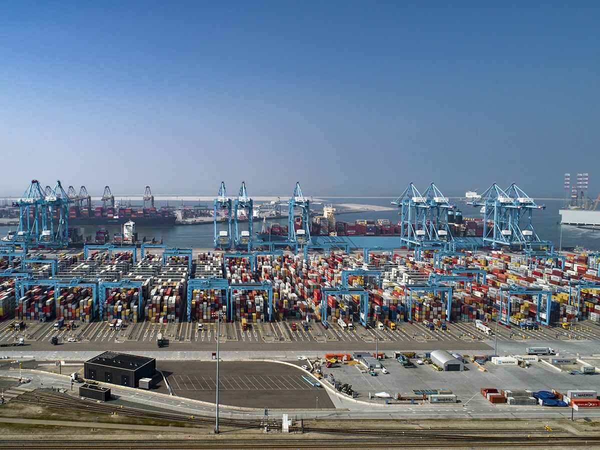 Luftbild eines geschäftigen Containerhafens mit unzähligen bunten Containern, großen blauen Kränen und Frachtschiffen im Wasser.