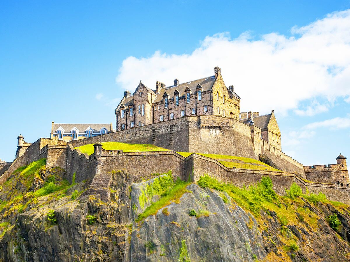 Edinburgh Castle auf Fels über grünen Hängen vor blauem Himmel. Historische schottische Festung mit Steinmauern.