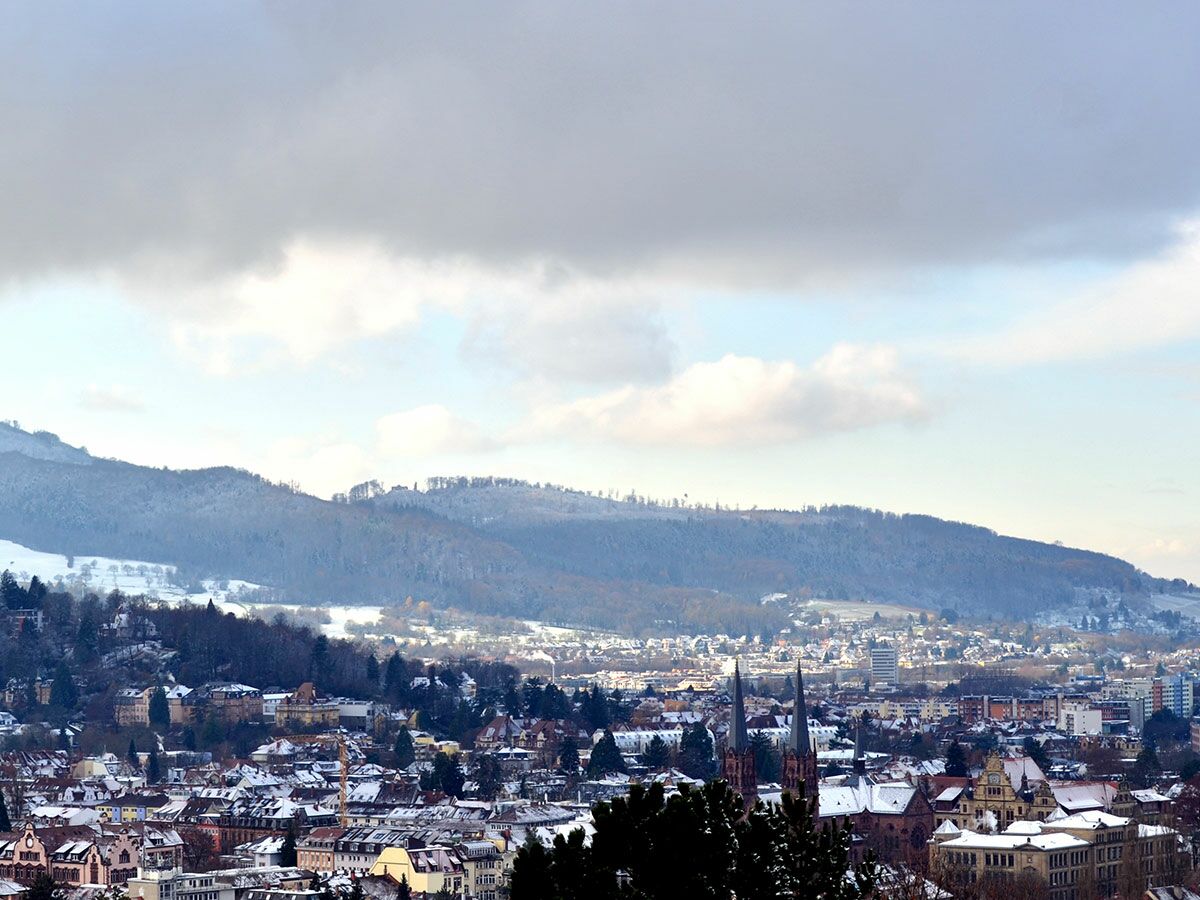Eine verschneite Winterlandschaft von Freiburg im Breisgau mit historischen Gebäuden, Kirchen und schneebedeckten Bergen im Hintergrund.