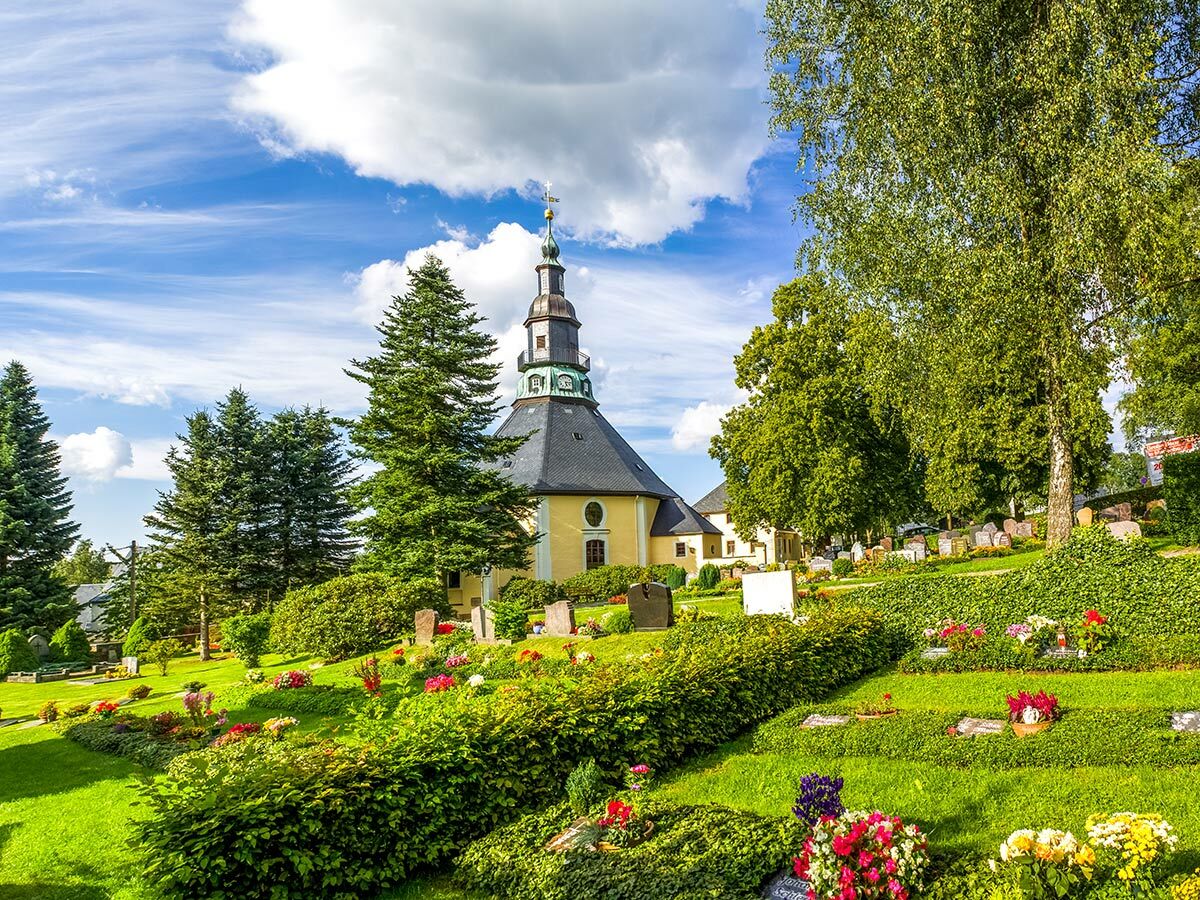 Idyllischer Friedhof mit einer Kirche, grünen Gräbern voller Blumen und Bäumen unter blauem Sommerhimmel.