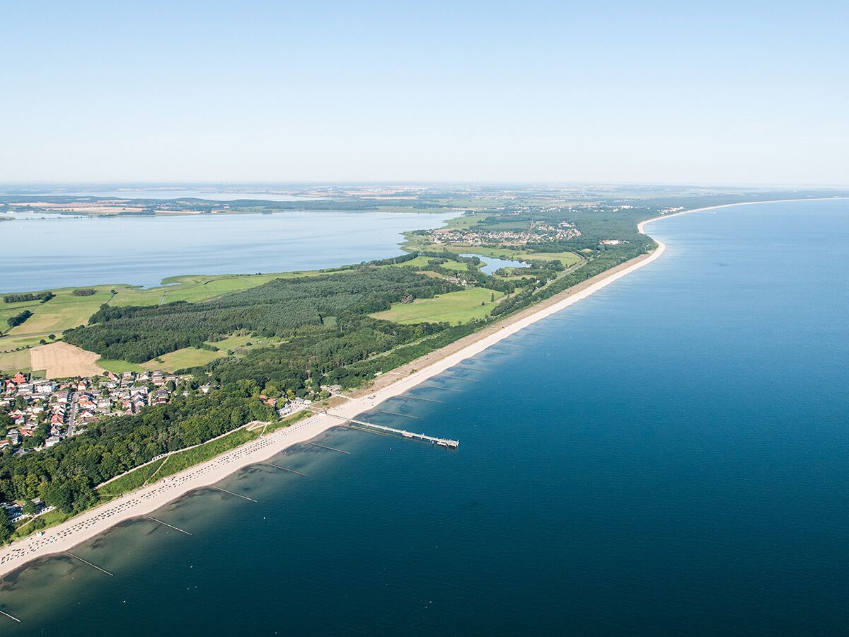 Luftaufnahme einer weiten Küstenlandschaft mit Sandstrand, blauem Meer, grünem Wald und einem Ort an einem Binnensee.