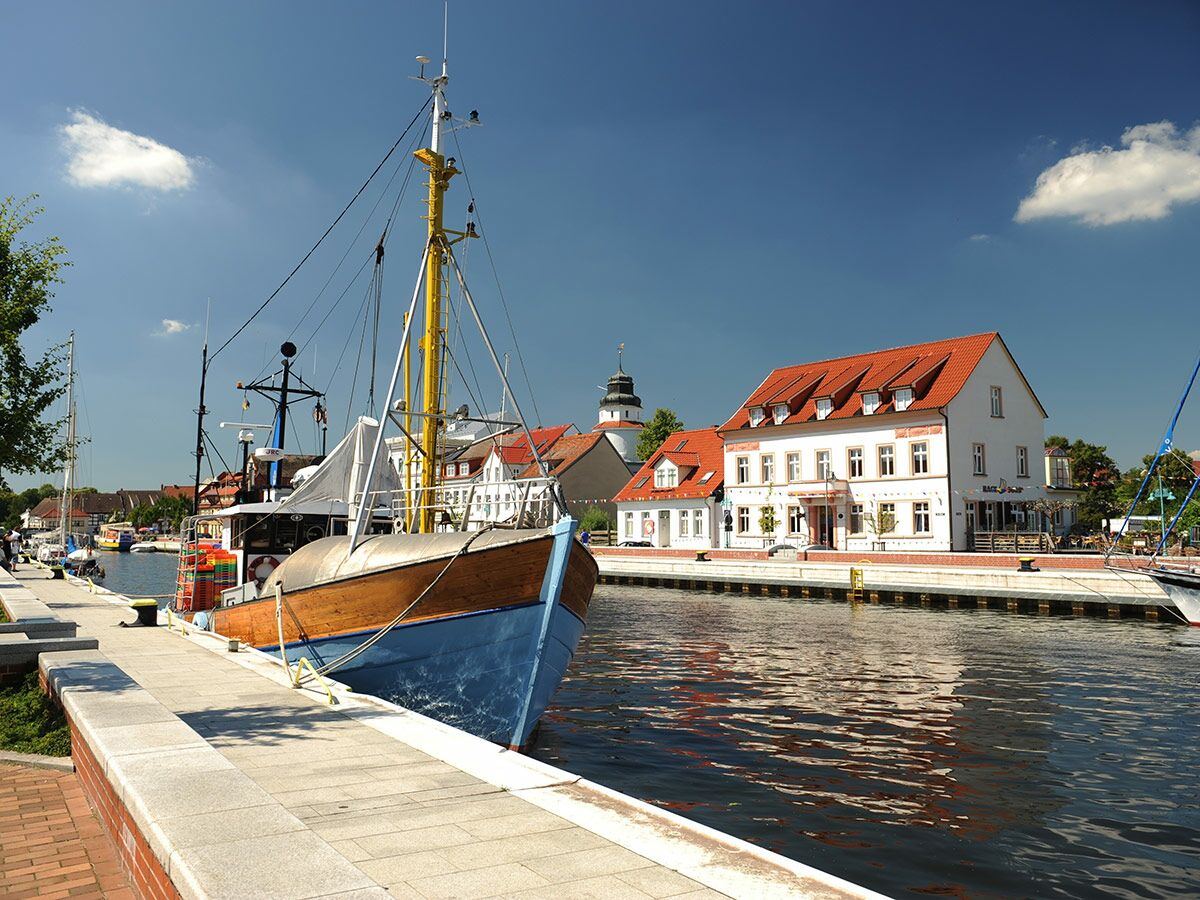 Malerischer Hafen mit Fischerboot, weißen Gebäuden mit roten Ziegeldächern, Kirchturm und blauem Himmel.