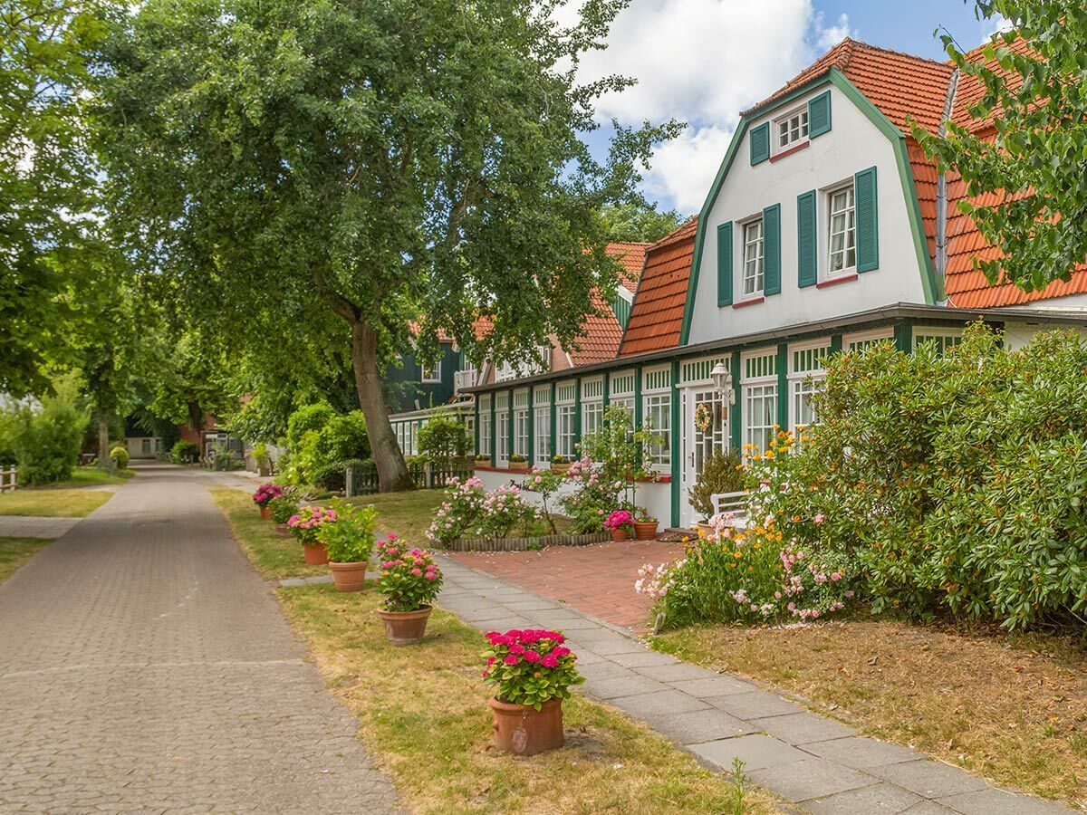 Traditionelles Wohnhaus mit rotem Ziegeldach, grünen Fensterläden, blühendem Garten und alten Bäumen an einem sonnigen Weg.