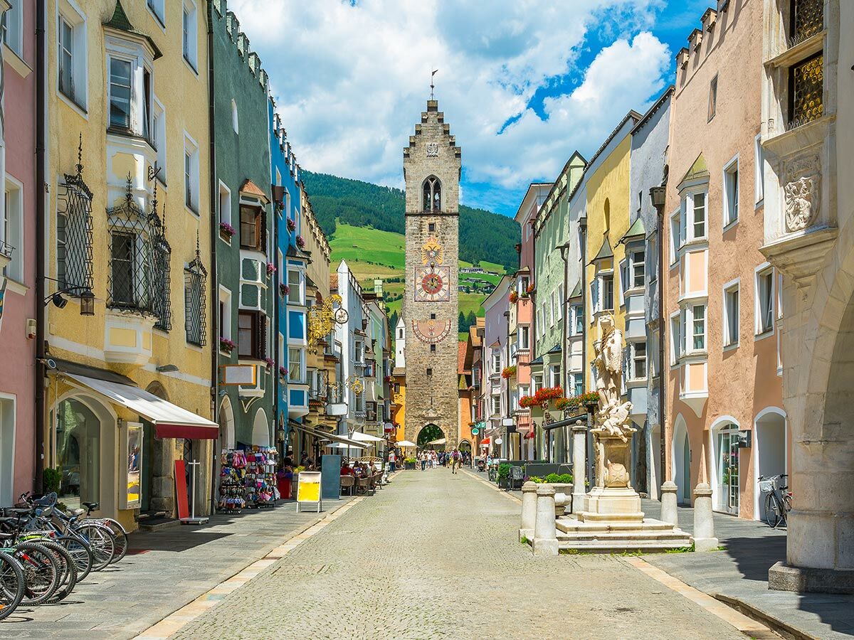 Malerische Altstadtgasse in Brixen, Südtirol, mit bunten Häusern, Weisser Turm, Brunnen und Bergkulisse unter blauem Himmel.